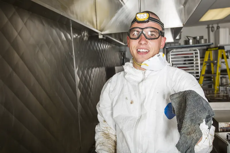 Technician in coveralls and goggles standing beside a stainless steel exhaust canopy in a commercial kitchen, ready to begin a hood cleaning visit