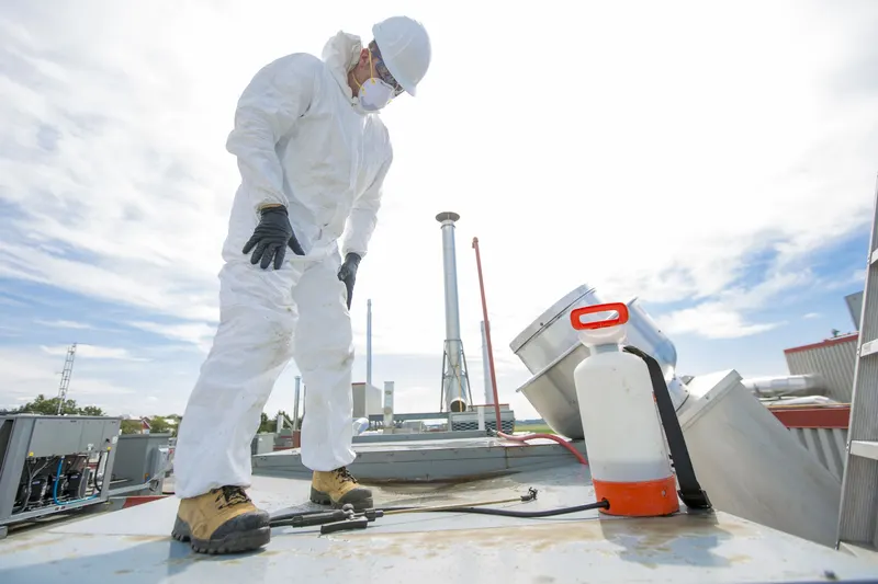 Technician in white coveralls, a white hard hat, a respirator and black gloves standing on a flat commercial roof beside an orange and white pump sprayer, looking down at a spray wand and hose on the roof surface, with an upblast exhaust fan and a rooftop unit behind him under a bright cloudy sky