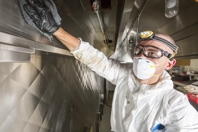 Technician in a white coverall, goggles and headlamp reaching up with a gloved hand to wipe the plenum wall inside a commercial kitchen exhaust hood