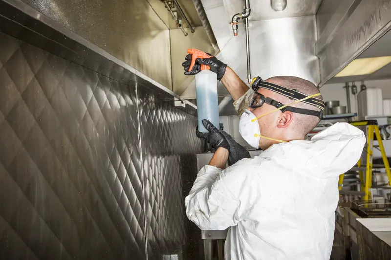 Technician in protective gear spraying degreaser across the underside of a stainless steel commercial kitchen canopy with the baffle filters removed