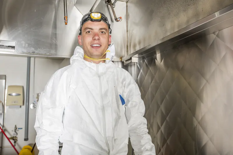 Technician in coveralls and a headlamp working beneath a stainless commercial kitchen hood canopy