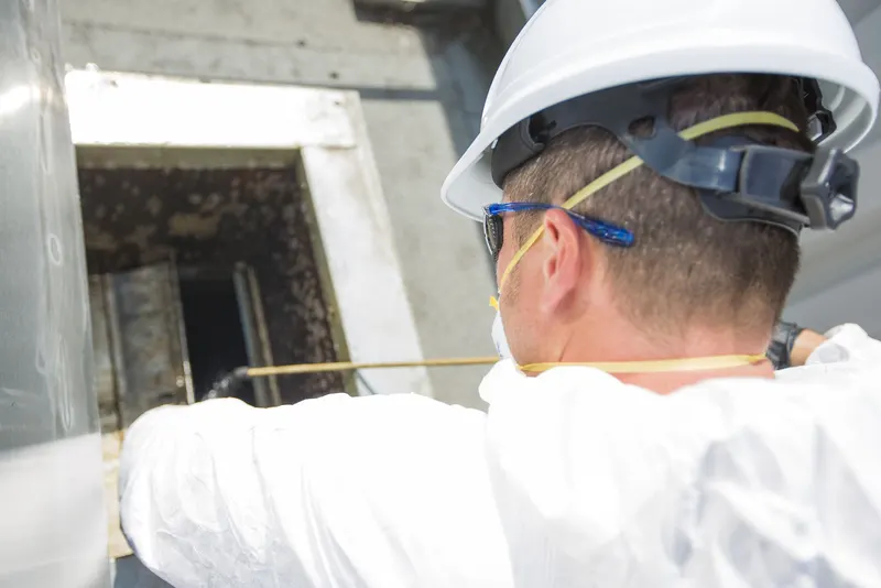 Worker in a white hard hat and safety glasses reaching into an open access panel on a grease-darkened commercial exhaust duct
