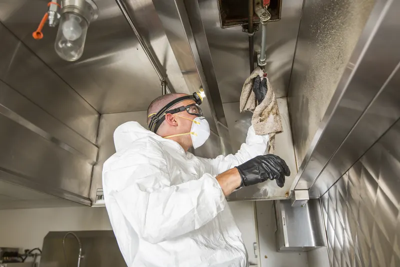 Technician in protective coverall and respirator wiping the underside edge of a stainless steel kitchen exhaust canopy with a cloth