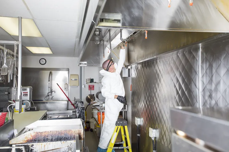 Hood cleaning technician on a step ladder wiping the front edge of a stainless steel exhaust canopy in a commercial kitchen with the appliances covered
