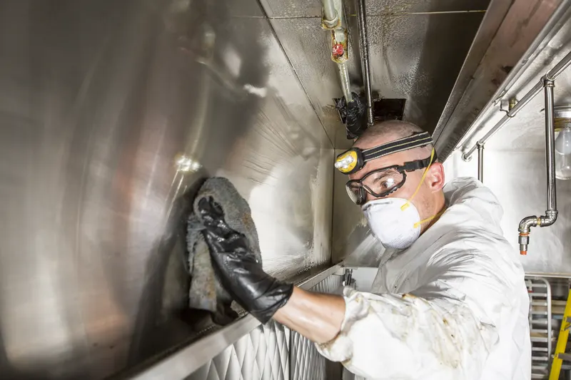 Technician in goggles and a respirator wiping grease from the polished inside wall of a kitchen exhaust duct with a gloved hand