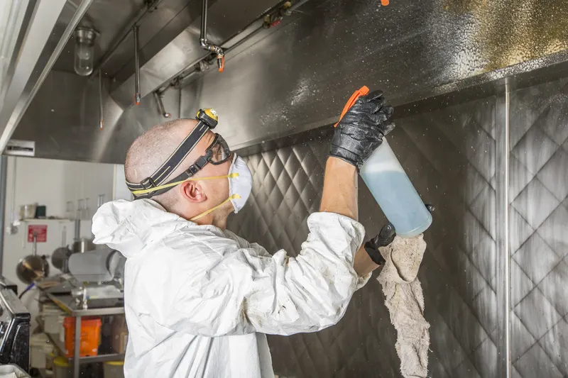 Technician in white coveralls, safety goggles, a respirator mask and a headlamp spraying blue degreaser from a bottle onto the underside of a stainless steel canopy hood while holding a rag, with a quilted stainless wall behind him in a commercial kitchen